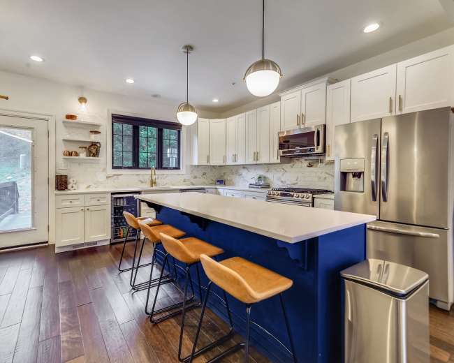 The image shows a modern kitchen featuring a blue island with bar stools, white cabinetry, stainless steel appliances, and large windows letting in natural light.
