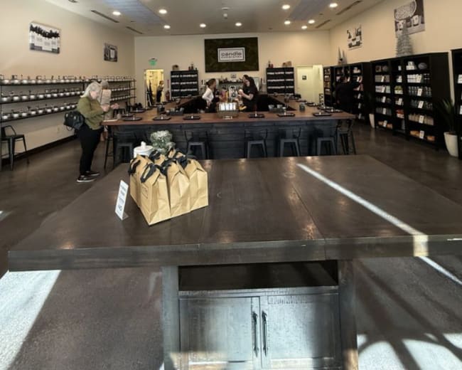 The image shows a spacious candle shop with black shelves lined with various candles and a central table where customers are interacting.