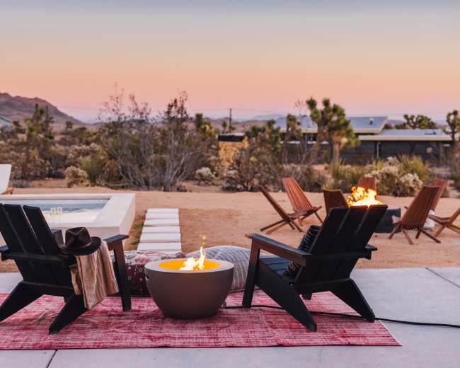 Two black Adirondack chairs face a circular fire pit, set on a red rug near a swimming pool with a desert landscape and mountain backdrop at sunset.