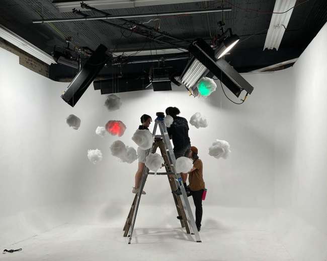 Three people are arranging decorative cloud props on a white studio set using a ladder.