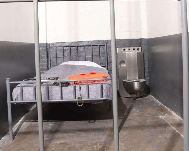 The image shows a sparse prison cell with a bed, a small table, and a toilet attached to the wall.