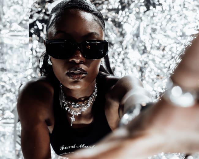 A person wearing sunglasses and jewelry crawls towards the camera against a reflective silver background.