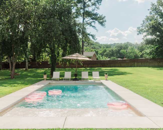 A swimming pool surrounded by green grass features pink inflatable floats and lounge chairs under a sun umbrella.