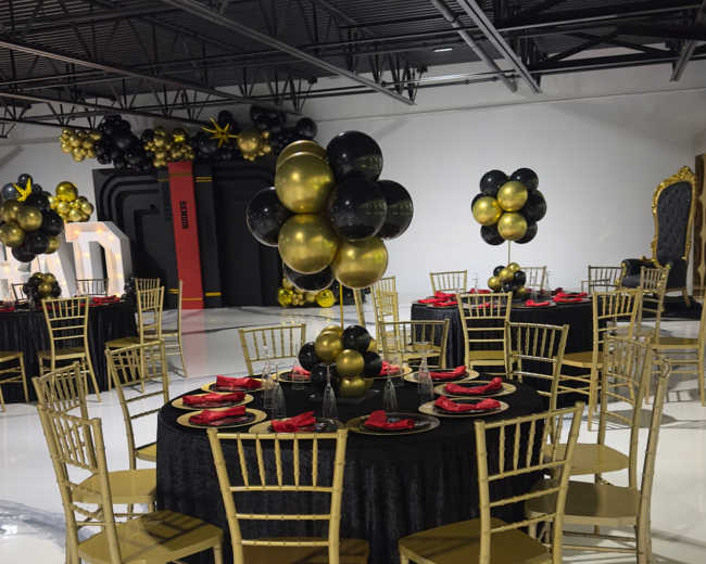 A banquet hall is set with round tables covered in black tablecloths, adorned with gold and black balloon centerpieces, and gold chairs arranged around each table.
