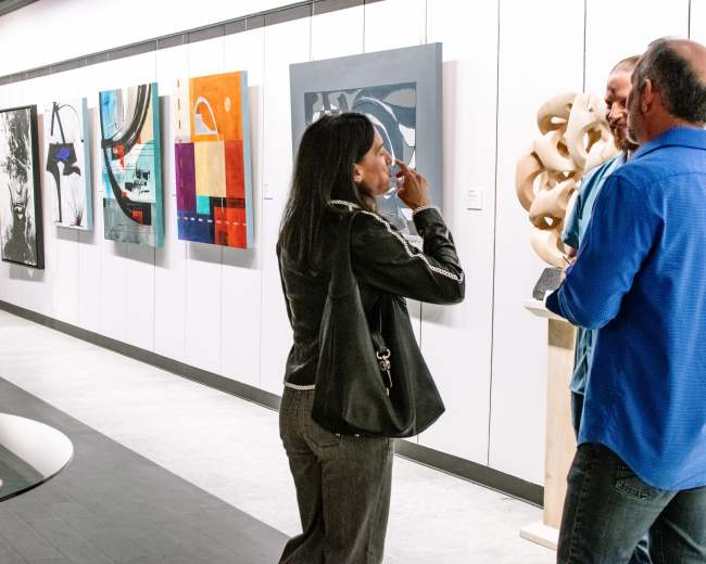 Two people converse in an art gallery featuring various paintings and a sculptural piece on display.