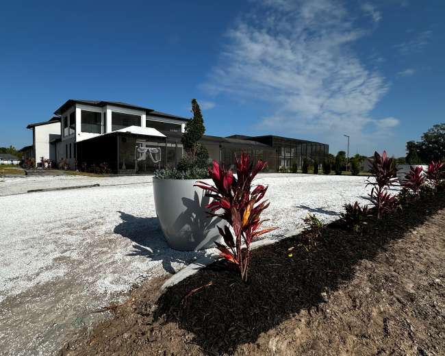 A modern two-story house with a garden area is flanked by colorful plants and a large decorative planter on a gravel path.