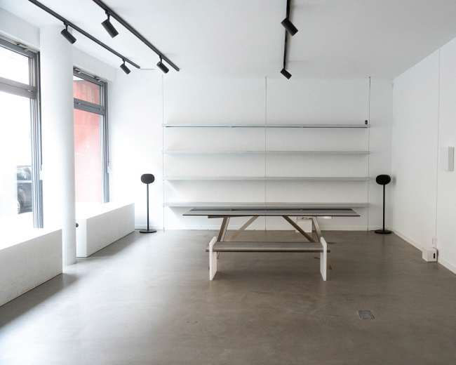 The image shows a minimalist interior space with a central table, two floor lamps, and empty shelves against white walls.
