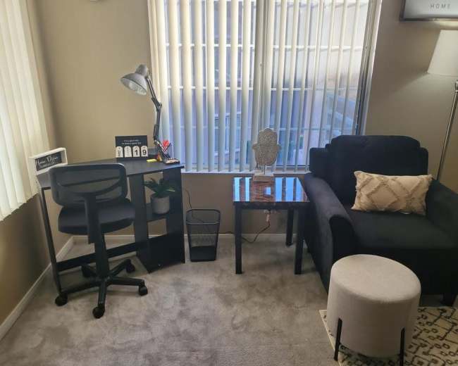 The living space features a black chair, a small table with a decorative item, a white ottoman, a desk with a lamp, and large windows letting in natural light.