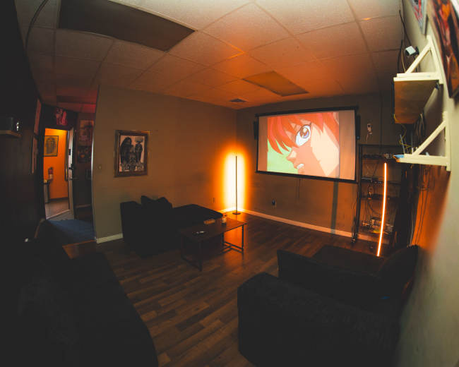 A cozy room features a projector screen displaying an animated character, with dark couches and ambient lighting.