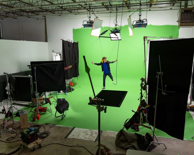 A person is suspended on wires in front of a green screen, surrounded by studio lighting and equipment.