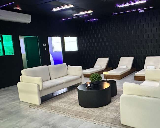 The image shows a modern relaxation room with two white sofas, a round black coffee table, and several lounge chairs arranged around a cozy area rug.