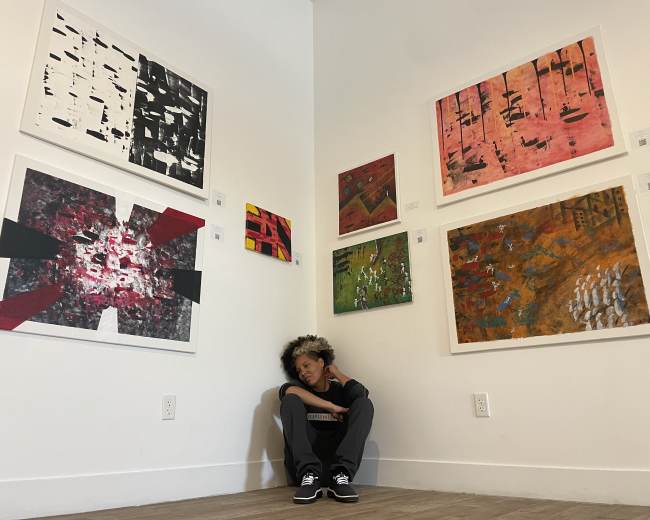 A person sits in a corner of an art gallery surrounded by various colorful paintings on the walls.