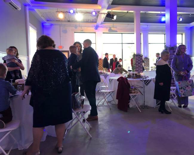 The image shows a spacious event venue with guests interacting and seated at white tables, while a few people stand around, surrounded by decorations and a large window letting in natural light.