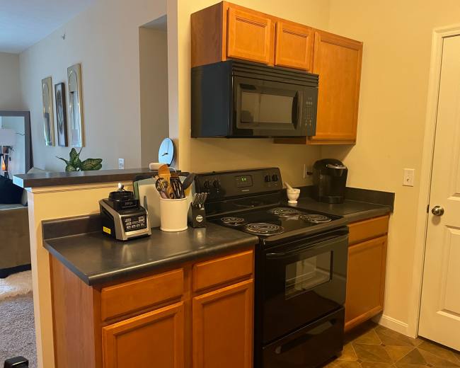 The image shows a kitchen area featuring wooden cabinets, a black microwave and stove, and various kitchen appliances on the countertop.