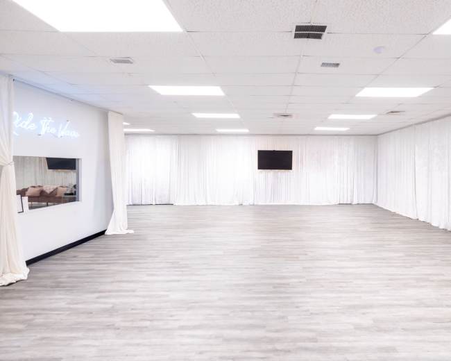 An empty room with light-colored wooden flooring, white drapes along the walls, and a television mounted on the back wall.