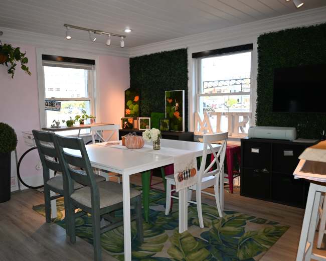 The image shows a bright, modern dining area with white tables and chairs, green plants on the walls, and a window with a view of an outdoor space.