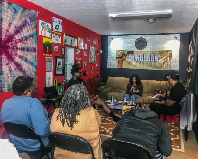 A group of people is seated in a cozy room decorated with vibrant artwork and banners, engaging in conversation around a small table.