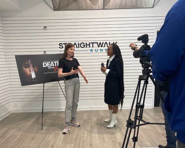 Two women are engaged in conversation in a retail space, with one standing beside a large sign displaying "DEATH TO P" and a camera crew filming the scene.