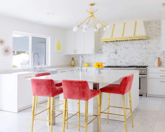 A modern kitchen features a large island with four red bar stools and a gold light fixture above, complemented by a marble backsplash and white cabinetry.