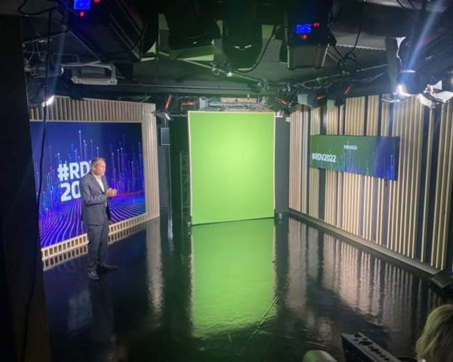 A man stands on a glossy black stage in a studio equipped with green screens and display monitors.
