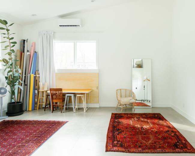 A spacious room features a wooden table with chairs, a large mirror, two area rugs, and light-colored walls, with exercise equipment stored in the corner.