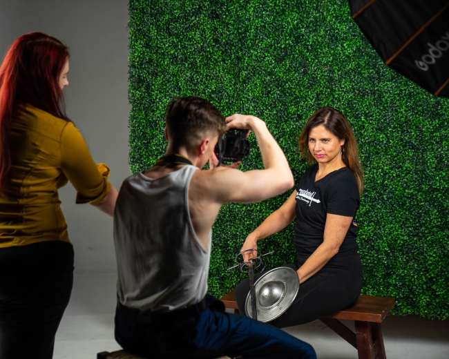 A photographer captures a woman posing with a silver bowl in front of a green backdrop while another woman observes the shoot.