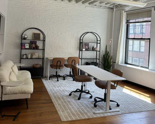 The image shows a modern office space with a white-brick wall, a light-colored rug, a long table with rolling chairs, and decorative shelving units.