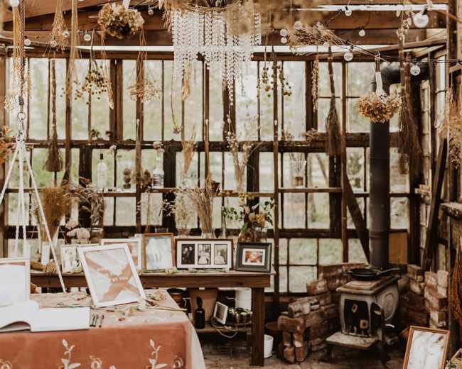 A rustic indoor space filled with hanging dried flowers and decorative elements, featuring a table with photographs in front of a windowed wall.