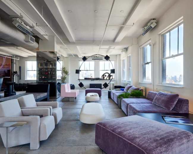 The image shows a modern, open-concept living space furnished with various colorful couches, a central table, and large windows providing natural light.