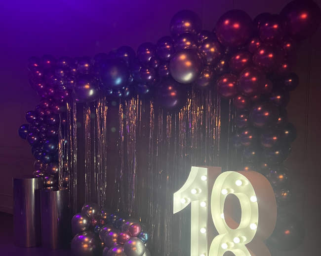 A decorative setup for an event, with a large illuminated "18" sign surrounded by a backdrop of metallic balloons and a curtain of shiny fringe.