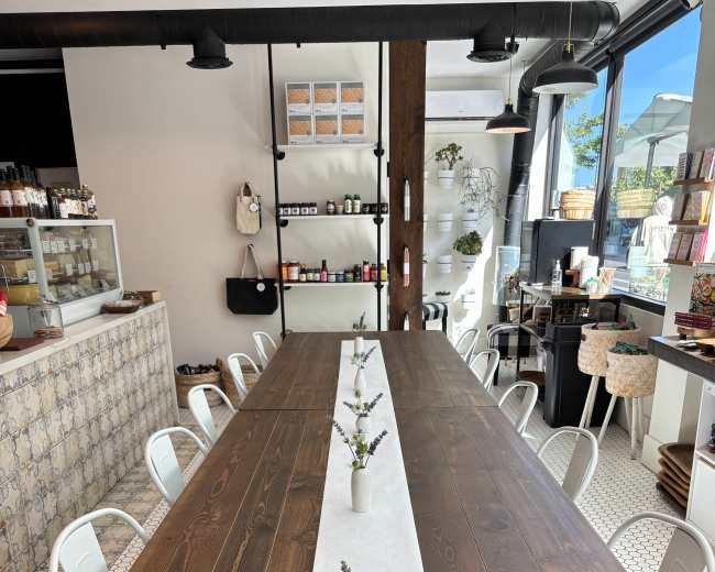 A long wooden table with a white runner and small vases sits in a café, surrounded by white chairs and featuring a tiled floor and a display counter in the background.