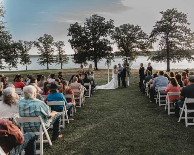 A couple stands under an arch exchanging vows in front of an audience seated on white chairs beside a body of water surrounded by trees.
