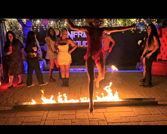 A performer in a pink outfit jumps over a line of fire while several spectators watch in a brightly lit party setting.