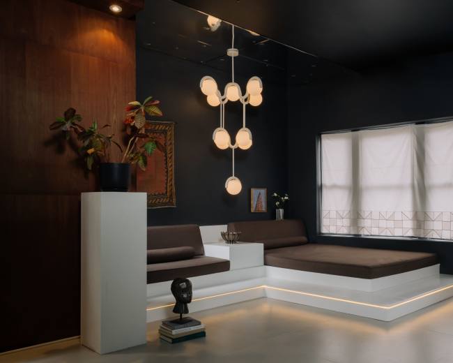 The image shows a modern interior living space featuring a low, sectional sofa, a sculptural light fixture, and a large window with light filtering through sheer curtains.