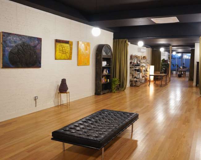 The image showcases a spacious art gallery with wooden flooring, featuring various paintings on the walls, a black lounge chair in the foreground, and shelves filled with items in the background.