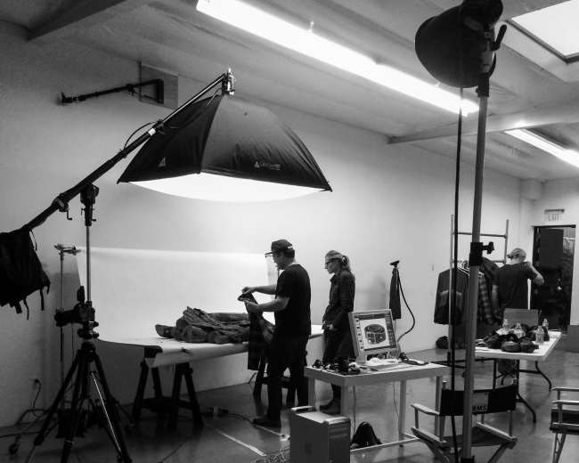 A photography studio is set up with a backdrop and various equipment, while individuals assist in capturing images of a subject on a table.