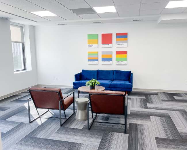 A modern office lounge features a blue sofa, two brown chairs, a round table, and colorful artwork on the wall.