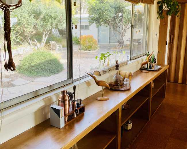 A wooden sideboard with various kitchen items and houseplants stands by a large window overlooking a landscaped garden.