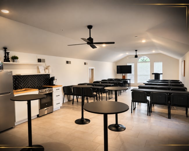 The image shows a modern meeting room featuring rows of chairs, a kitchenette area, and a podium under a high ceiling.