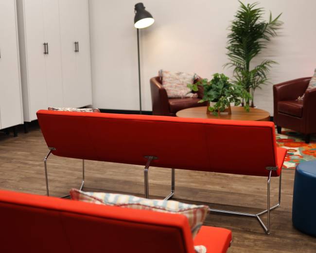 The scene features a modern lounge area with red seating, a round table, and a potted plant, set against a backdrop of white cabinets and a floor lamp.