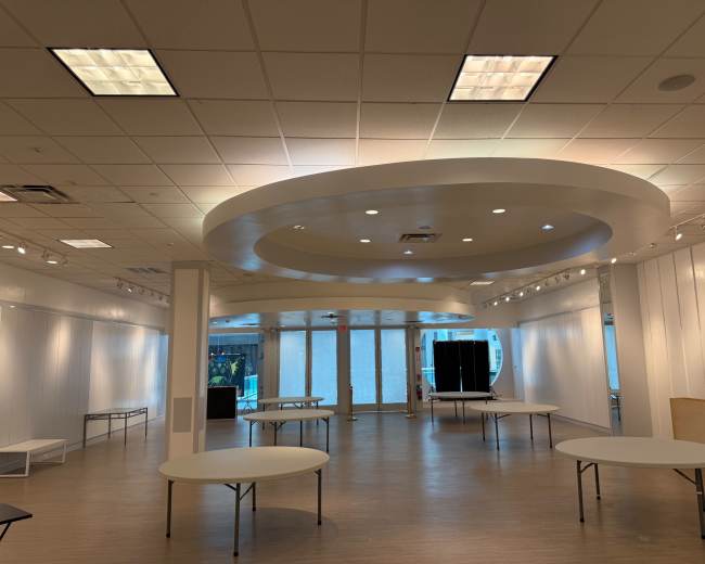 The image shows a spacious room with white walls, multiple round tables set up, and a ceiling featuring circular designs and recessed lighting.