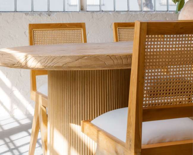A round wooden table with two caned chairs is positioned near large windows adorned with natural light, along with a vase of flowers on the table.