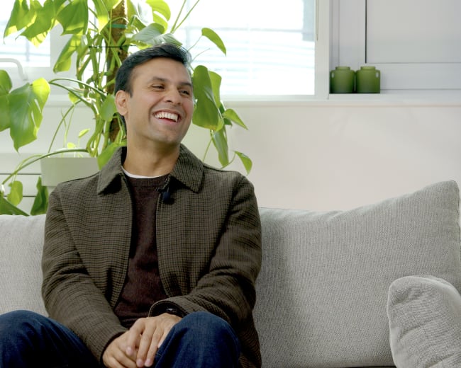 A man sits on a grey sofa with a green plant in the background, smiling while engaged in conversation.