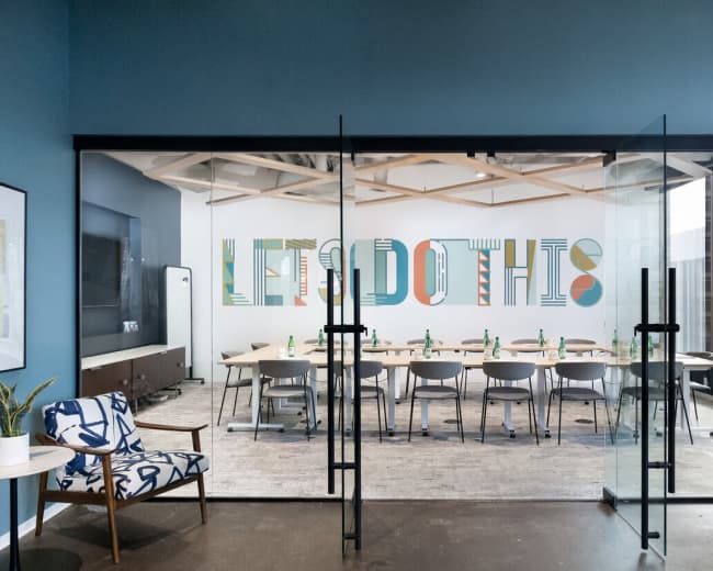 A modern conference room features large glass doors, a colorful motivational wall design, and a rectangular table surrounded by chairs.