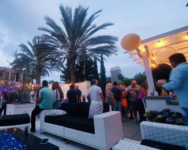 A crowd gathers at an outdoor evening party surrounded by palm trees and decorative lighting.