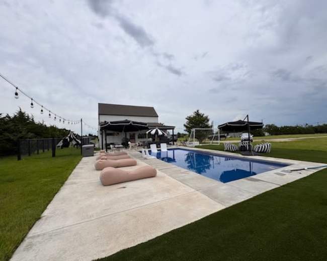 A rectangular pool surrounded by lounge chairs and a grassy area, with a building in the background under an overcast sky.