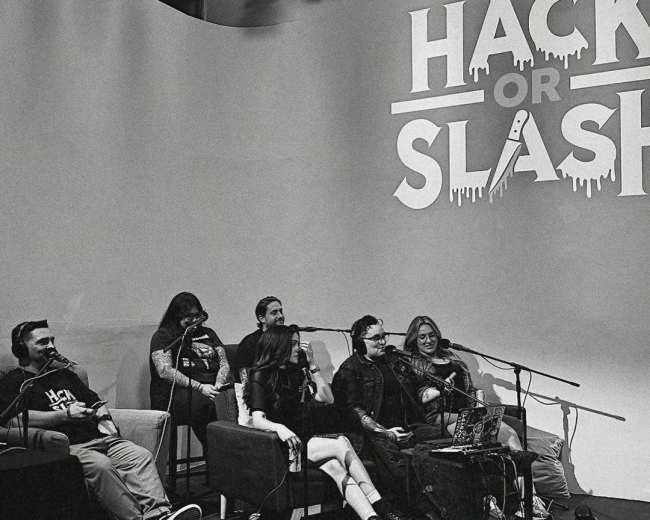 A group of five people sits at a table in front of a large backdrop that reads "HACK OR SLASH," with various microphones and equipment displayed.