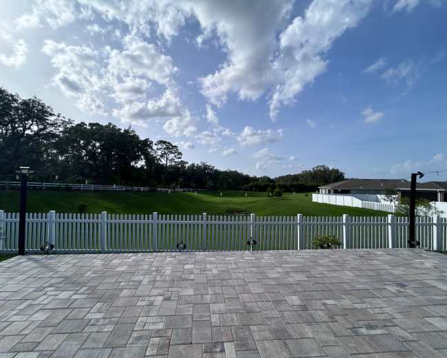 The image shows a spacious backyard with a lawn, bordered by a white fence under a partly cloudy sky.