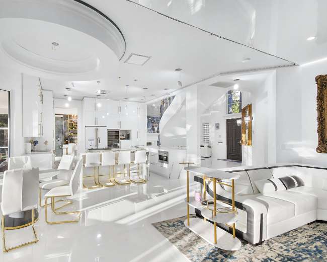 A modern kitchen and living area with sleek white furnishings, gold accents, and large windows allowing natural light.