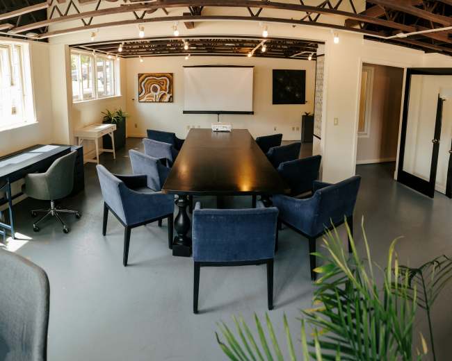 The image shows a modern conference room with a large table surrounded by upholstered chairs, white walls, and artwork on the walls.